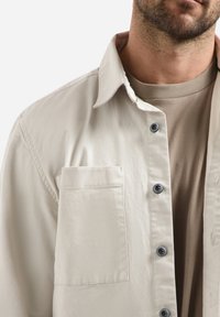 Lichtbeige shirtjasje gemaakt van zachte stof, met een zak, een kraag en donkere knopen voor het sluiting. Daaronder zit een beige top.