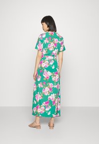 Vestido maxi floral em verde com flores rosas e brancas. Apresenta mangas curtas e um cinto atado na cintura. Textura de tecido suave.