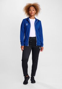 Giacca sportiva blu con cappuccio, zip bianca e logo; abbinata a pantaloni da jogging neri con strisce laterali e vita elasticizzata; scarpe nere.