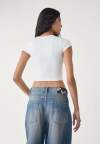Witte, kortlopende, crop top gecombineerd met hoge, lichtblauwe denimjeans met een zwart merklabel op de tailleband.
