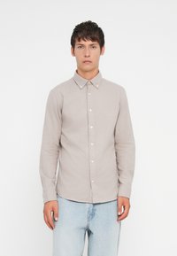 Košela s dlhým rukávom v svetlobéžovej látke s golierom na gombíky, so bielymi gombíkmi a rovnou spodnou časťou. Nosená s svetlomodrými džínsami.