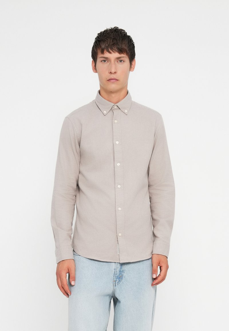 Košela s dlhým rukávom v svetlobéžovej látke s golierom na gombíky, so bielymi gombíkmi a rovnou spodnou časťou. Nosená s svetlomodrými džínsami.