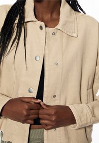Beige spijkerjack met grote kraag, zilveren drukknopen en twee voorzakken. De stof heeft een zachte textuur en een iets losse pasvorm.