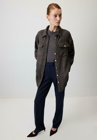 Brązowa marynarka w jodełkę z złotymi guzikami, noszona na szarym topie, zestawiona z granatowymi spodniami i ciemnymi szpilkami z ostro zakończonym noskiem.