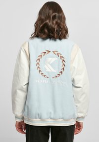 Modrá a krémová bombera s vyšitým logom, textúrovanou tkaninou, rebrovanými manžetami a pások. Na zadnej strane sa nachádza dizajn vavrínovej koruny.