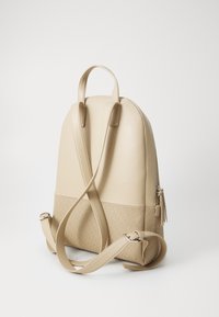 Zaino in pelle beige con forma sagomata, spallacci regolabili e sezione inferiore texturizzata con motivo circolare. Dettaglio della tasca con zip incluso.