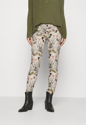 Slim-fit beige pants with botanical and bird print paired with black ankle boots and an olive green long-sleeve top on a plain background.