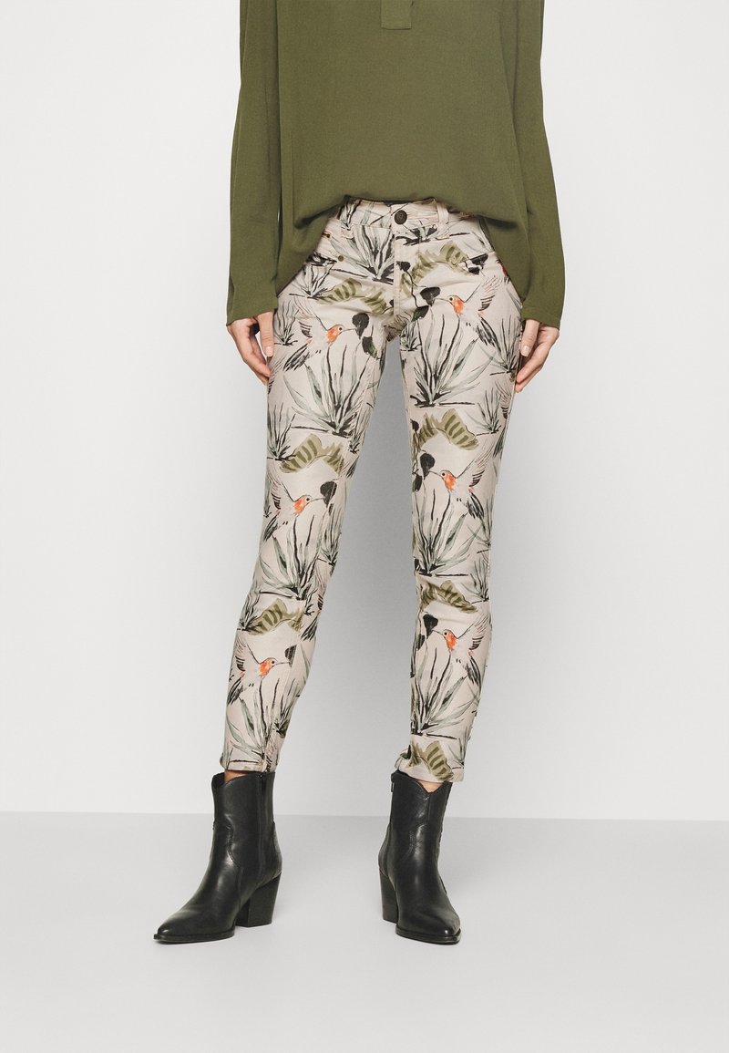 Pantalon beige slim fit avec imprimé botanique et oiseaux, associé à des bottines noires et un haut à manches longues vert olive sur un fond uni.