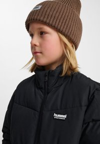 Svart, vadderad jacka med hög krage och dragkedja, med en liten vit Hummel sportklädeslogotyp. Brun ribbad mössa på huvudet.
