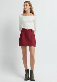 Witte geribbelde lange mouwen top, met een off-the-shoulder ontwerp. Rode gestructureerde A-lijn rok met een voorste knoop, gecombineerd met olijfgroene enkellaarsjes.