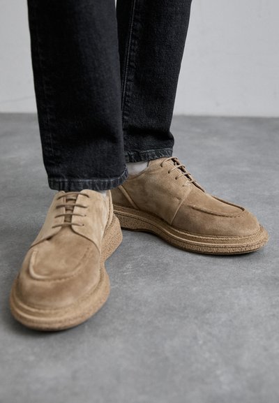 Beige ruskindssko med en glat tekstur og rund tå. Udstyret med snørebånd, en tyk tekstureret sål og suppleret med mørk denimbukser.