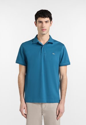 Giovane uomo con capelli corti e ricci che indossa una polo a maniche corte blu teal e pantaloni beige, in piedi davanti a uno sfondo bianco semplice.