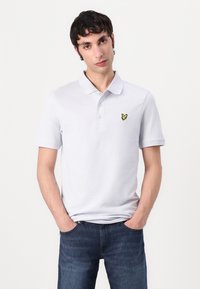 Polohemd aus leichtem grauem Baumwollstoff, mit einem gelben gestickten Logo auf der linken Brust, kombiniert mit dunkelblauen Jeans. Kurze Ärmel, klassischer Kragen.