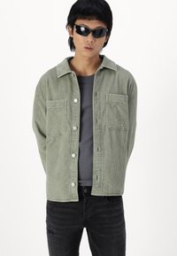 Lys grønn kordfløyelsjakke med knappelukking, to brystlommer og klassisk krage, brukt over en grå skjorte og svarte jeans.