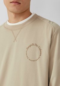 Beige sweatshirt met een ribgebreide ronde halslijn. Beschikt over een cirkelvormige print met de tekst "Escape To The Unknown" en "PUSHING BOUNDARIES." Zachte textuur.