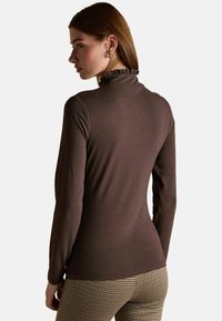 Pruun pika varrukaga kõrge kaelusega topp, millel on volangkrae, sujuv tekstuur ja figuuri rõhutav lõige, koos houndstooth-mustriga pükstega.