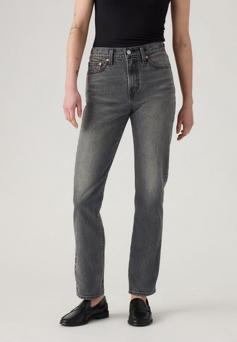 Personne portant un jean droit noir délavé taille haute, un haut noir sans manches et des mocassins noirs, debout devant un fond uni.