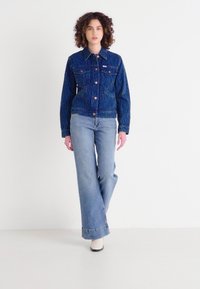 Chaqueta de mezclilla azul acolchada con dos bolsillos en el pecho y botones naranjas, combinada con jeans acampanados de un azul claro y botas de tacón blancas.