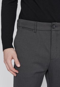 Man med mörkgrå randiga byxor och svart långärmad topp, hand avslappnad vid vänster lår.