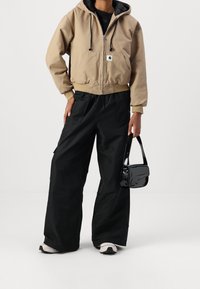 Beige hoodie met ritssluiting en een voorzak, zwarte wijde broek en een kleine zwarte lakleren handtas. Sport schoenen zijn ook zichtbaar.