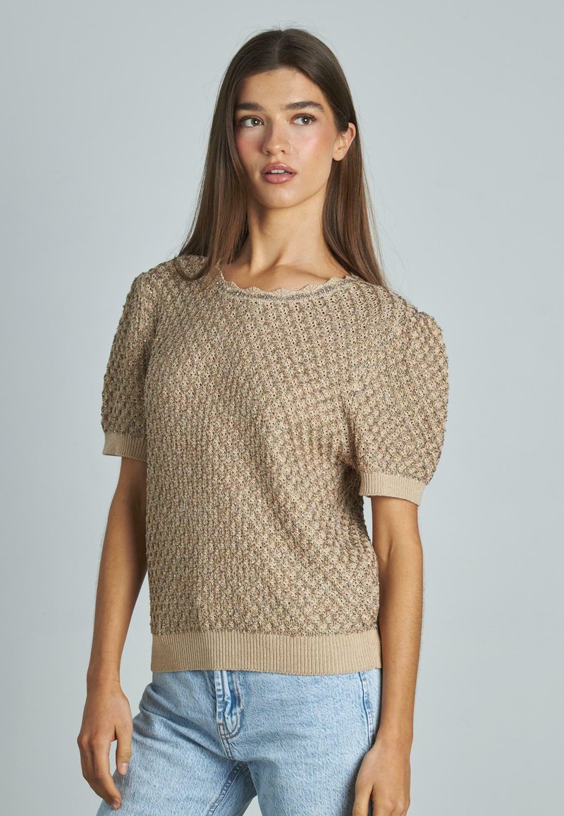 Maglione lavorato a maglia beige con motivo testurizzato, maniche a puff corte e polsini a costine. Collo a scallop e vestibilità morbida. Abbinato a jeans blu chiaro.