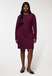 Abito lungo bordeaux a maniche lunghe con silhouette aderente e cintura coordinata. Tessuto liscio, orlo fino al ginocchio, indossato con scarpe con tacco nere.