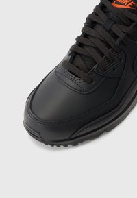 Tenis cu piele neagră, cu șireturi negre, accente texturate pe cutia degetelor și o talpă din cauciuc. Prezintă un mic logo portocaliu pe limbă.