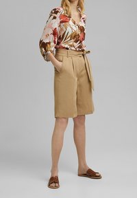 Blusa floreale con maniche a 3/4, caratterizzata da tonalità di rosa, rosso e marrone. Pantaloni corti beige a vita alta con tasche e cintura. Sandali marroni.