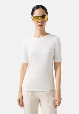 Kvinde iført hvid kortærmet top og cremefarvede bukser, med glat, mørkt hår trukket tilbage, der modellerer gule sportsbriller med toning.