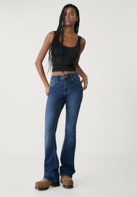 Blaue Schlagjeans mit einem Fünf-Taschen-Design und leichter Naht. Kombiniert mit einem schwarzen Tanktop und braunen Clogs, die einen lässigen Stil zeigen.