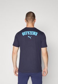 Tumši zila kokvilnas T-kreklā uz muguras ir teksts "CITYZENS" gaiši zilos burtos un neliels Puma logo zem tā. Īsas piedurknes un apaļa kakla izgriezums.