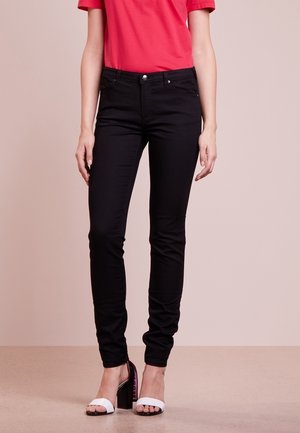 Femme portant un pantalon noir ajusté, une chemise rouge et des sandales blanches à talons hauts et ouverts à l'avant, debout sur un fond beige uni.