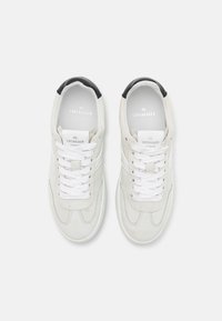 Białe zamszowe trampki o miękkiej fakturze, z czarnymi akcentami na pięcie, białymi sznurowadłami i markowymi wkładkami. Klasyczny sportowy design.