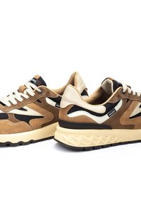 Zapatillas de ante marrón con acentos en negro y crema, superposiciones texturizadas, parte superior con cordones y suela con un patrón geométrico.