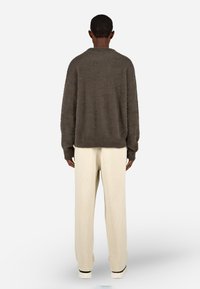 Osoba stojąca plecami, ubrana w puszysty, ciemnobrązowy sweter z długim rękawem, kremowe teksturowane spodnie i białe sneakersy na białym tle.