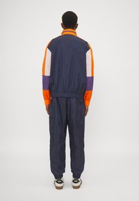 Marine joggedress med oransje og hvite fargeblokker, elastiske mansjetter og midjebånd, laget av lett, glinsende materiale.