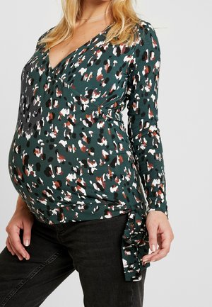 Mujer embarazada con una blusa de manga larga con estampado animal multicolor con lazo lateral, combinada con pantalones negros sobre un fondo liso.