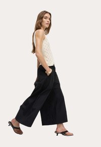 Vitt ribbat linne med guldprickar, matchat med vida svarta byxor. Modell som bär svarta flip-flops, stående mot en ljus bakgrund.