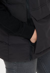 Sort puffervest med lynlåslukning, prydet med en sidelomme og en glat stofoverflade, båret over en sort langærmet skjorte.