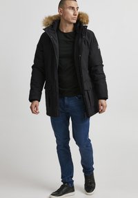 Sort parka-jakke med en hætte, der er foret med pels, lynlås foran, to forlommer og ribstrikkede manchetter, parret med blå jeans og sorte sneakers.