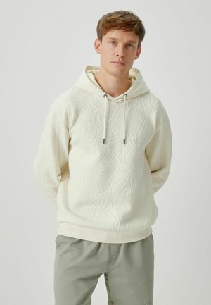 Young man wearing a textured off-white hoodie with drawstrings and light gray pants, standing against a plain white background.