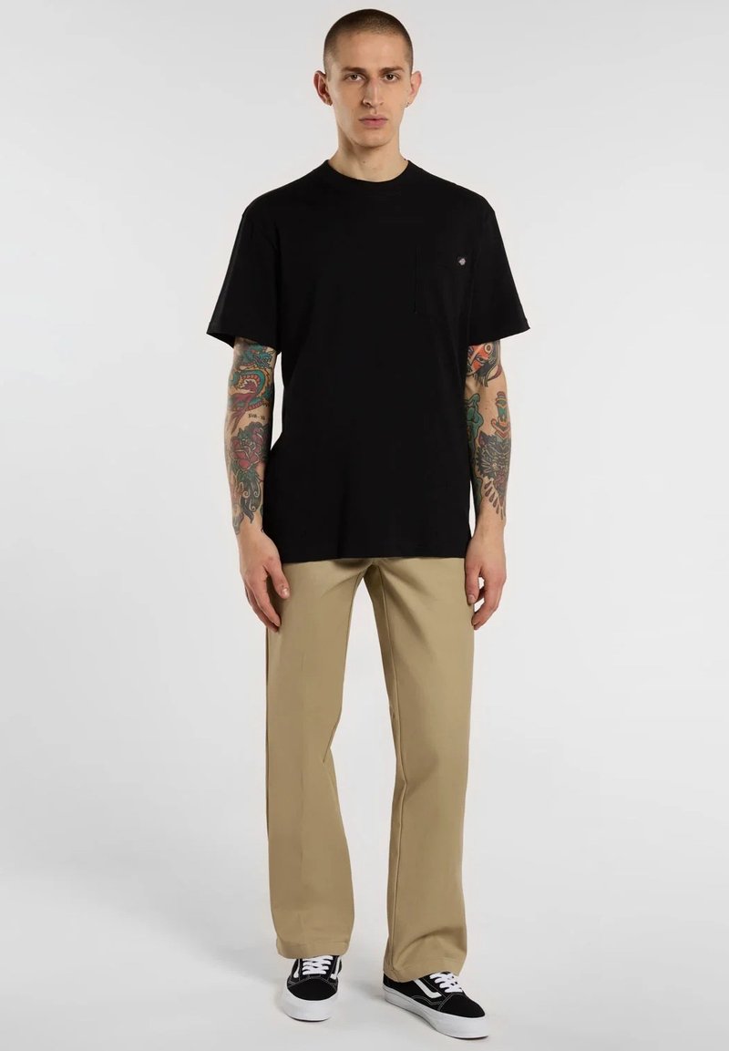 T-shirt nera in cotone con scollo tondo e taschino sul petto, abbinata a pantaloni beige a gamba dritta e sneakers nere.