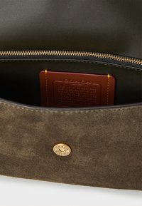 Olive suede interior of a handbag, featuring a gold zipper, embossed leather authenticity tag, and a metallic logo accent.