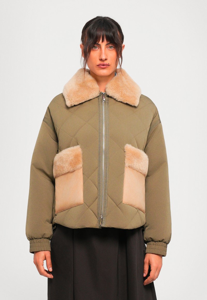 Donna che indossa una giacca trapunta verde oliva con colletto in pelliccia sintetica beige e tasche, in piedi contro uno sfondo bianco semplice.