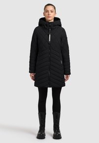 Zwarte gewatteerde pufferjas met capuchon, voorzien van een ritssluiting aan de voorkant, chevronpatroon en ritssluitingen aan de zijzakken, gecombineerd met zwarte leggings en laarzen.