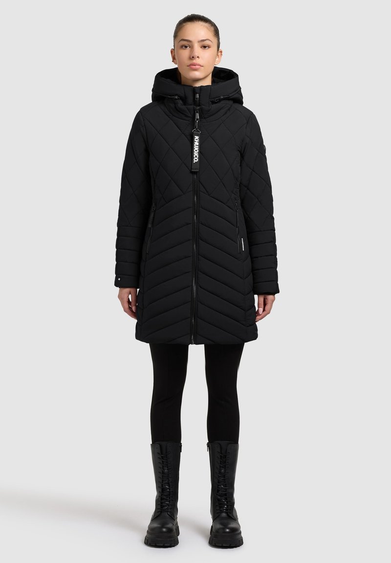 Zwarte gewatteerde pufferjas met capuchon, voorzien van een ritssluiting aan de voorkant, chevronpatroon en ritssluitingen aan de zijzakken, gecombineerd met zwarte leggings en laarzen.