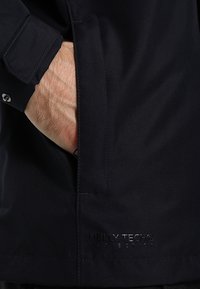 Main d'homme dans la poche d'une veste imperméable noire Helly Tech, montrant le poignet de la manche et le logo près de l'ourlet de la poche.