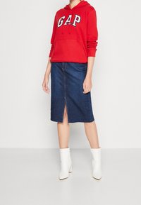 Sudadera roja con logo "GAP", falda de mezclilla con abertura frontal, combinada con botas de tobillo de tacón blanco. Texturas suaves, estilo casual.