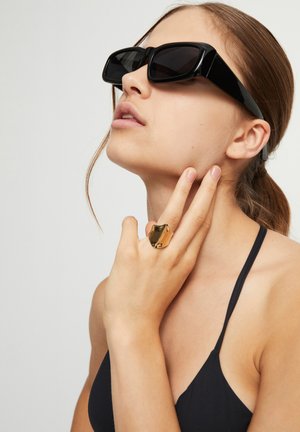 Gafas de sol rectangulares negras con brazos anchos, combinadas con un anillo dorado texturizado en la mano, sobre un fondo neutro.