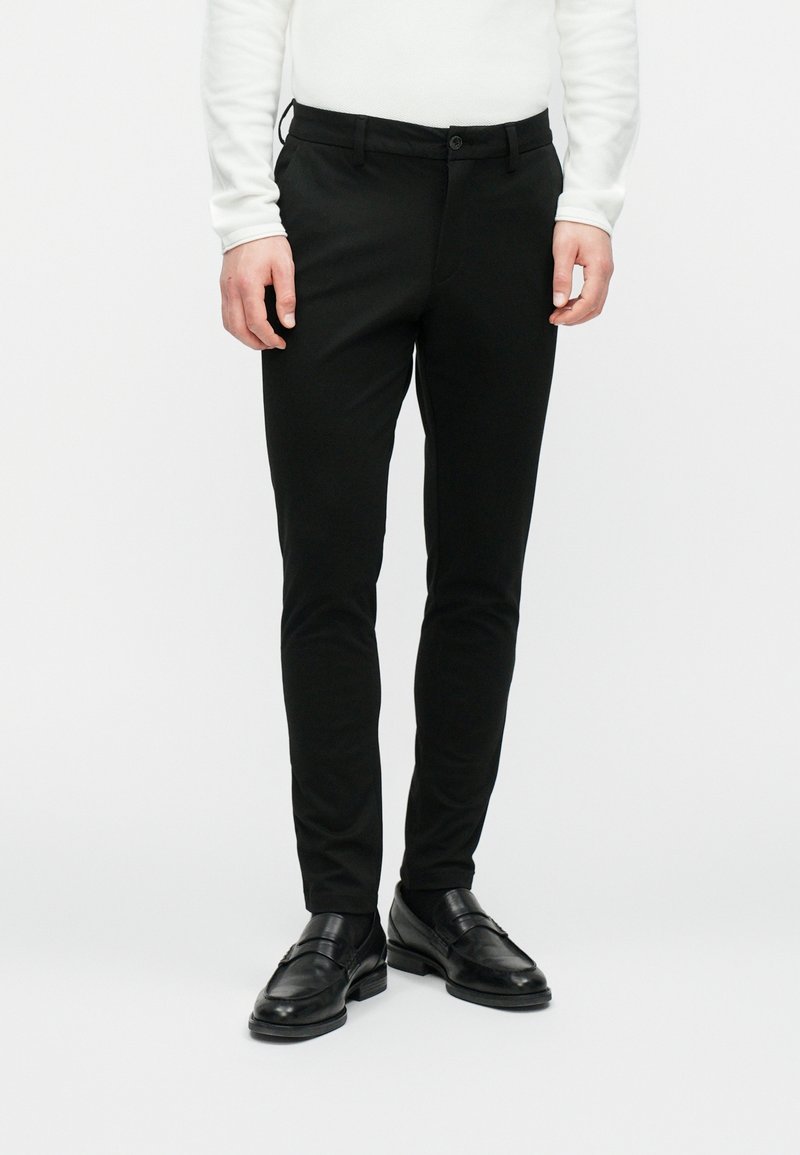 Homme portant un pantalon noir cintré, des mocassins en cuir noir et une chemise blanche à manches longues, debout sur un fond blanc.